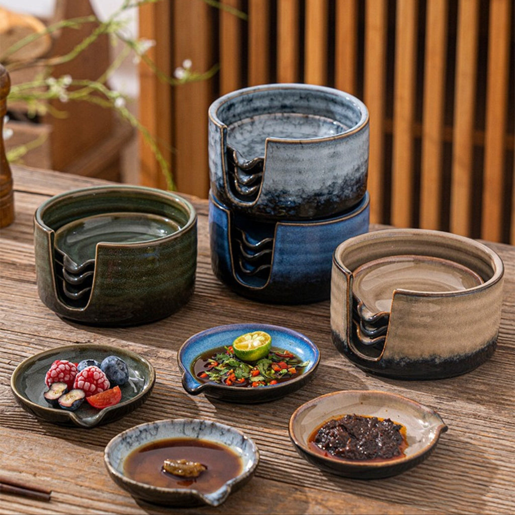 A set of four handmade ceramic dipping dishes arranged on a wooden table in a styled kitchen setting. The collection includes a dark green bowl, a stacked blue and black bowl, a beige bowl, and several small dipping plates with food, under the text 'Four Accompanying Dishes Collection'.