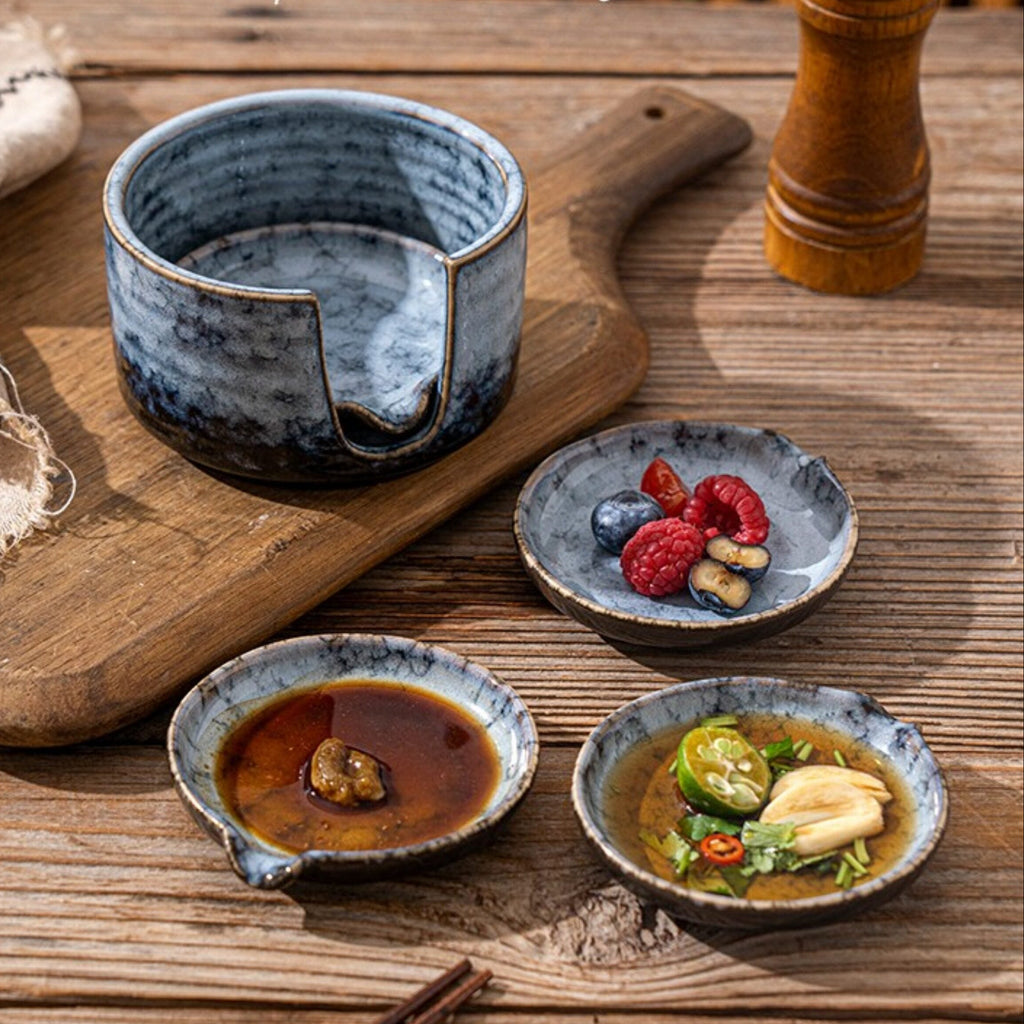 A ceramic tea pouring bowl set displayed on a wooden table, featuring a large blue-gray bowl with a pouring notch and three matching small dipping bowls with food, alongside a wooden pepper mill and a cutting board.
