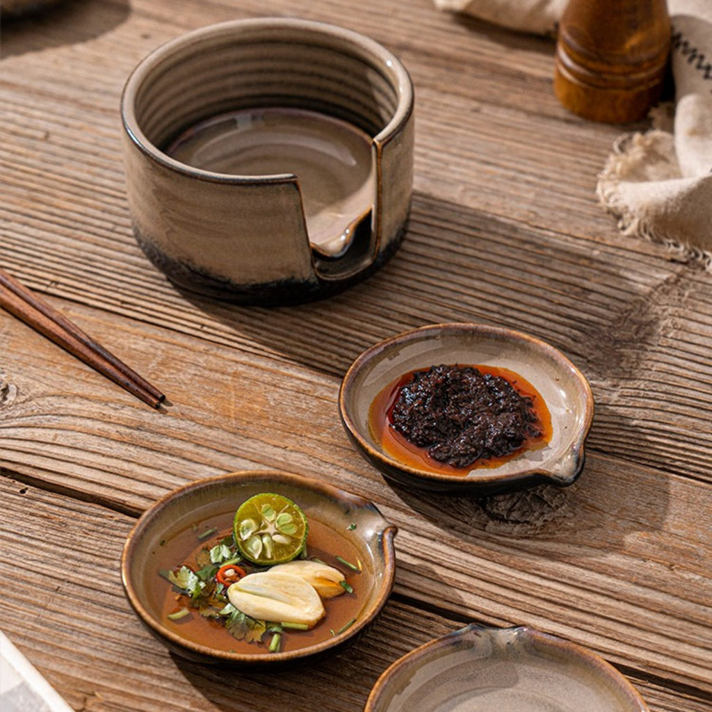A rustic ceramic set of small shallow bowls and a dipping bowl arranged on a wooden table, with chopsticks and food props around them.