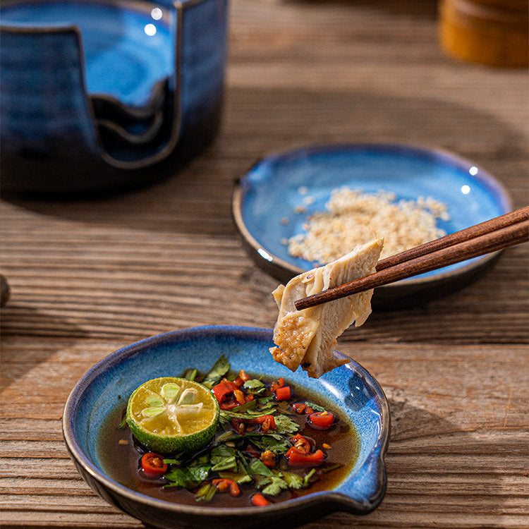 A ceramic dipping bowl with chopsticks lifting food, shown on a wooden table with other blue dishes and a lime slice. 