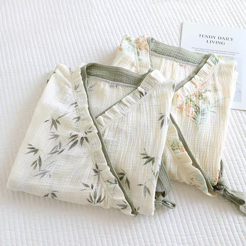 Two folded cotton muslin robes with ruffled trim, one in sage green and the other in olive green, both featuring a delicate floral and leaf print. They are styled on a white quilted bedspread next to a card that reads 'FUNDY DAILY LIVING'.