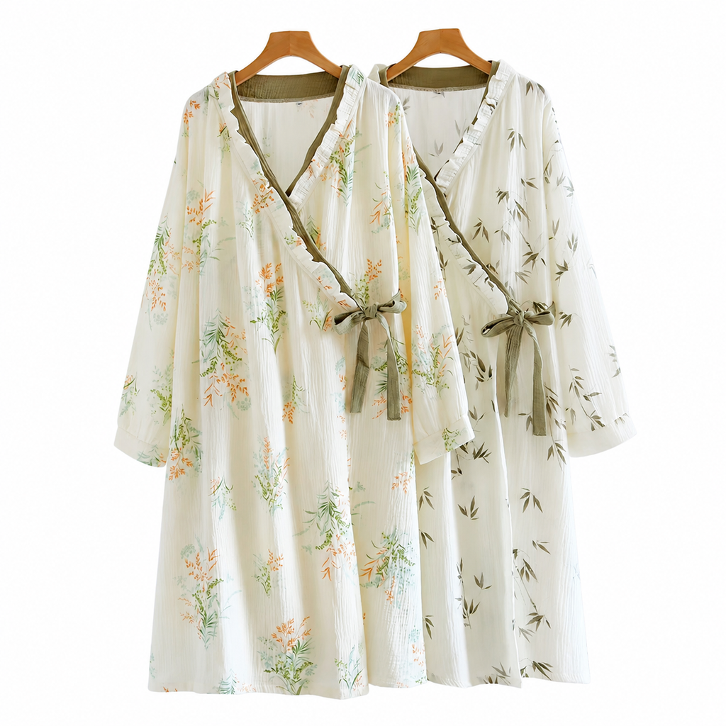 Two lightweight wrap-style dresses hanging on wooden hangers against a plain white background. The dress on the left has a cream base with orange floral and green botanical prints, while the dress on the right has a similar cream base with a green bamboo print.