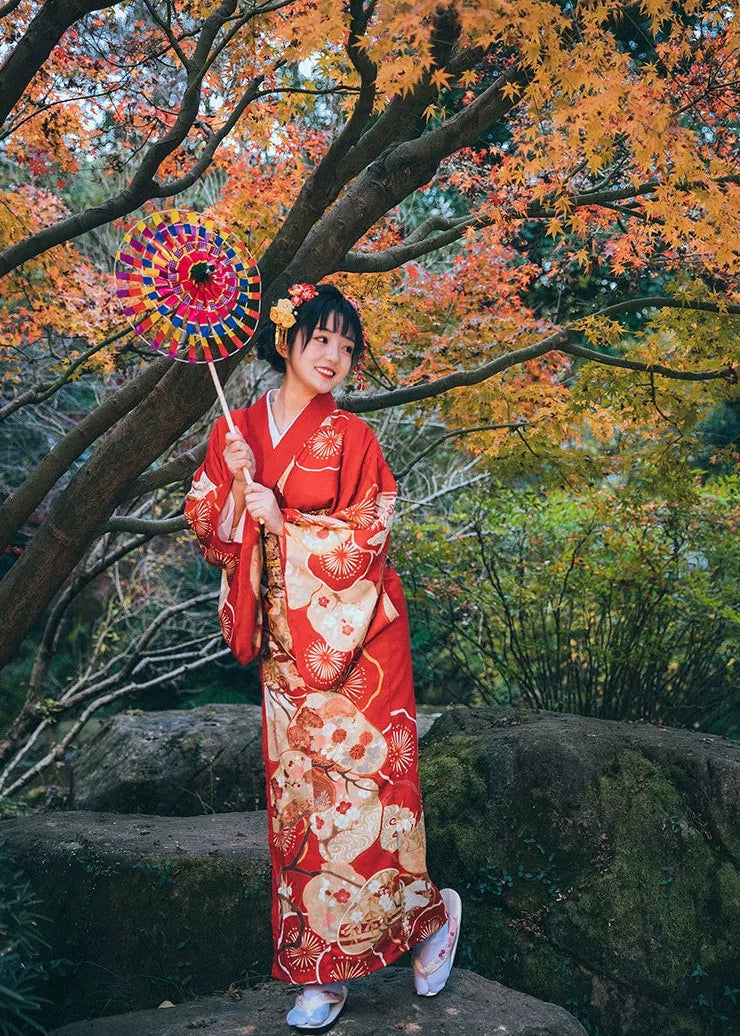 Woman in a red kimono holding a colorful umbrella in an autumn setting with trees and rocks.