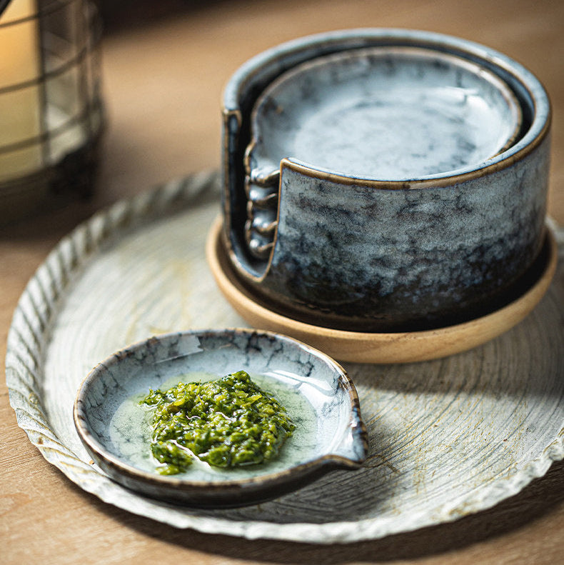A set of handmade ceramic tableware, including a small leaf-shaped dish with green condiment, a stack of bowls, and a shallow bowl, arranged on a round tray on a wooden table.